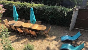 Um pátio externo com mesa e cadeiras de madeira, cercado por arbustos e um viveiro.