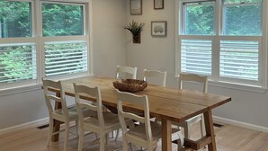 A dining room with a wooden table and white chairs, large windows with blinds, and a small plant on the wall.