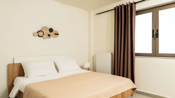 A bedroom with a wooden bed, white bedding, a wall-mounted light, a radiator, and a window with curtains. - Elysee apartment 1 (Athens)