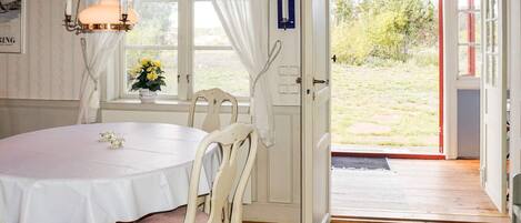 Una habitación con una mesa redonda, sillas blancas, una lámpara colgante, una ventana con cortinas y una puerta que da al exterior.