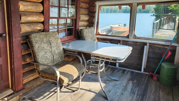 Una cabaña con interior de madera, una mesa con sillas, un ventanal con vistas a un lago y un muelle, y una puerta.