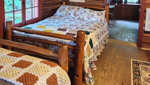 Un dormitorio de cabaña de troncos con una cama de madera, un colcha, un banco de madera y vista a la cocina a través de una puerta abierta.