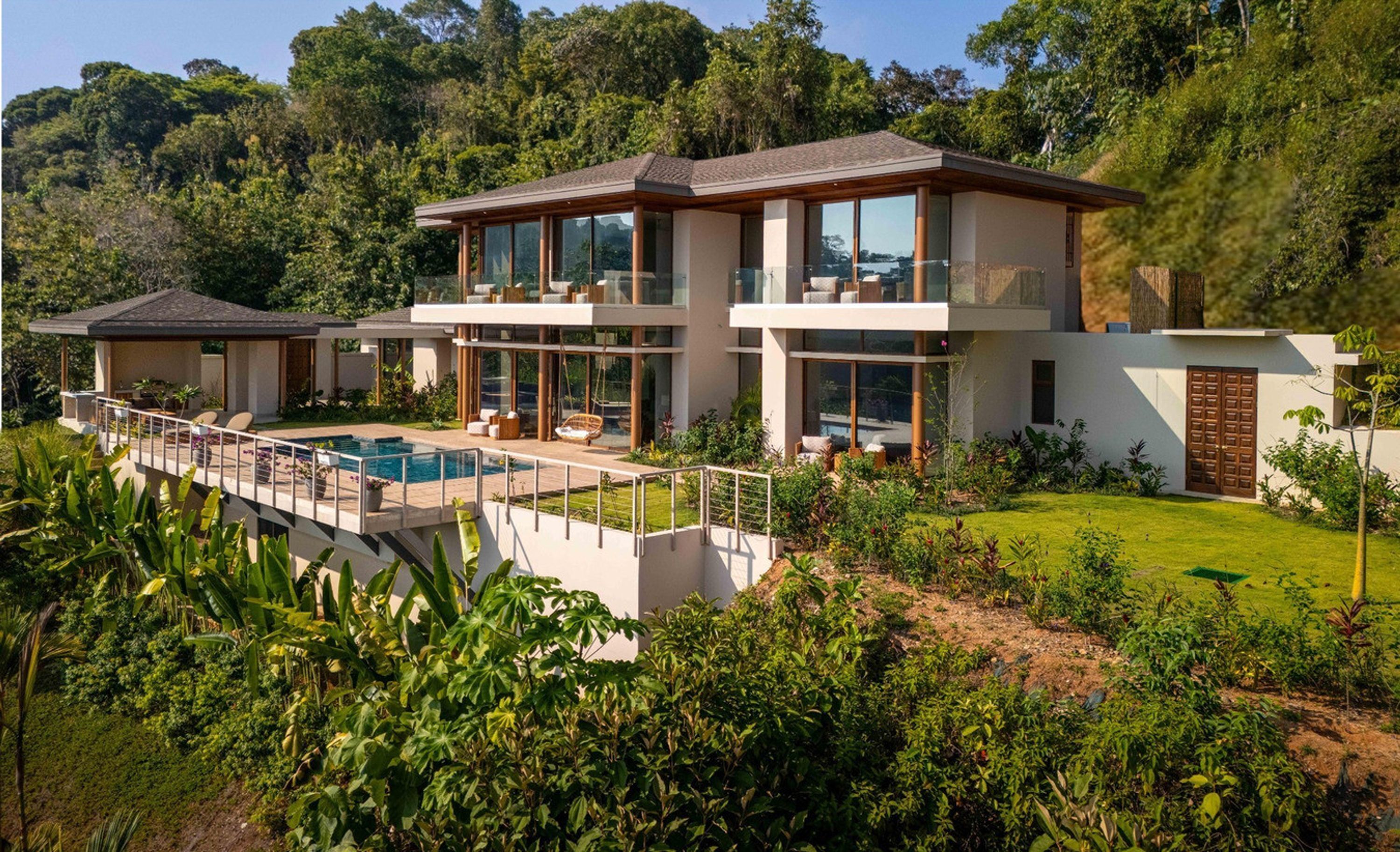 A modern two-story house with a large glass facade, a swimming pool, and lush greenery surrounding the property.