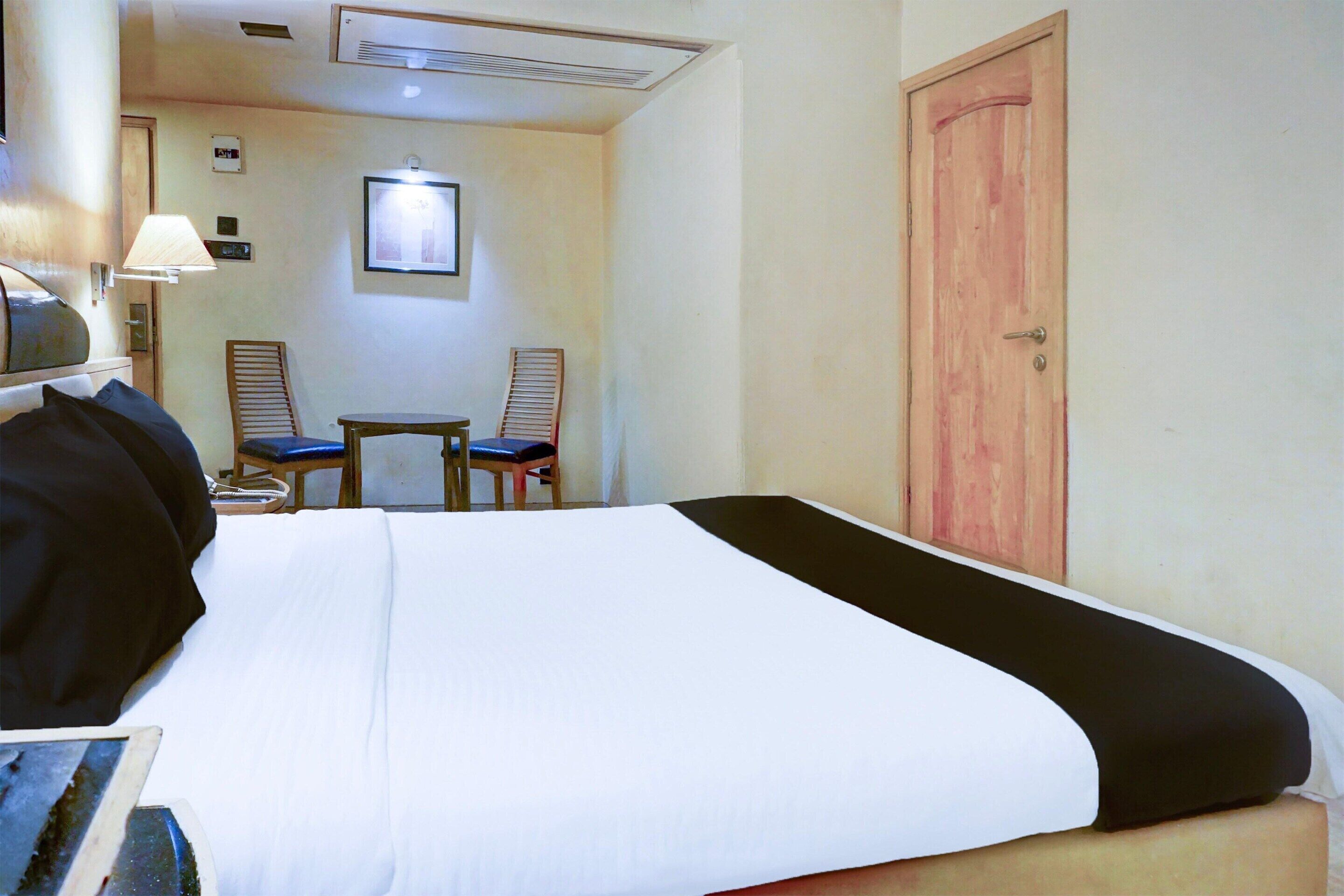 A hotel room with a bed, a desk, two chairs, a wooden door, and a wall-mounted picture.