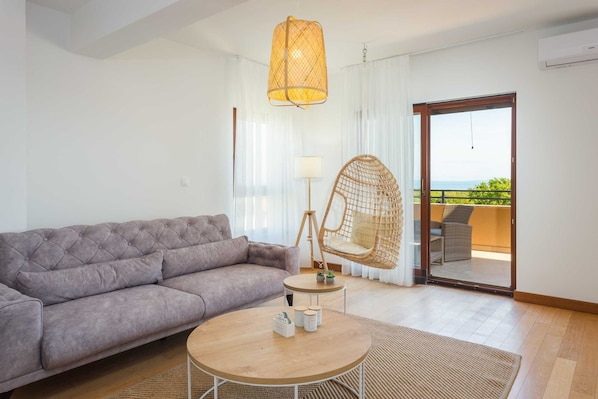 A modern living room with a grey sofa, a round wooden coffee table, a wicker chair, and a view through a sliding glass door to an outdoor balcony.