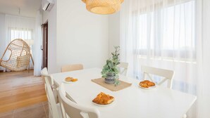 A dining area with a white table, chairs, a woven hanging light fixture, and a potted plant.