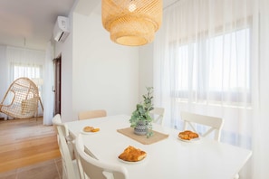 Un coin repas avec une table blanche, des chaises, un luminaire suspendu en fibres tressées et une plante en pot.