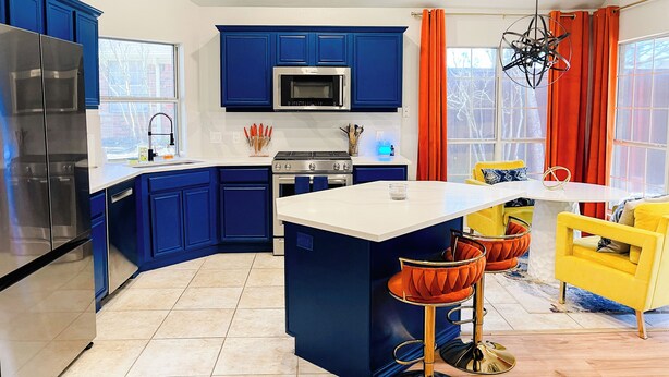 A modern kitchen with blue cabinets, a stainless steel refrigerator, a microwave, and a stove. There is a dining area with a yellow chair and a table. - Radiant Retreat with fitness room (McKinney)