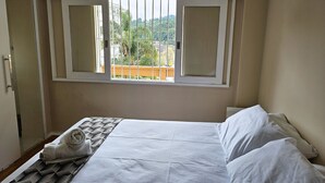 Un dormitorio con una cama, una ventana con contraventanas blancas, vistas al verdor exterior y una puerta a la izquierda.