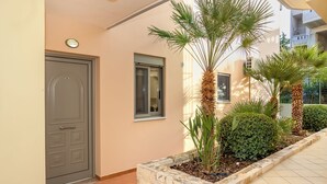 A building entrance with a grey door, a small window, and a stone planter with plants. - Casa Kamara I1 (Kounoupidiana)