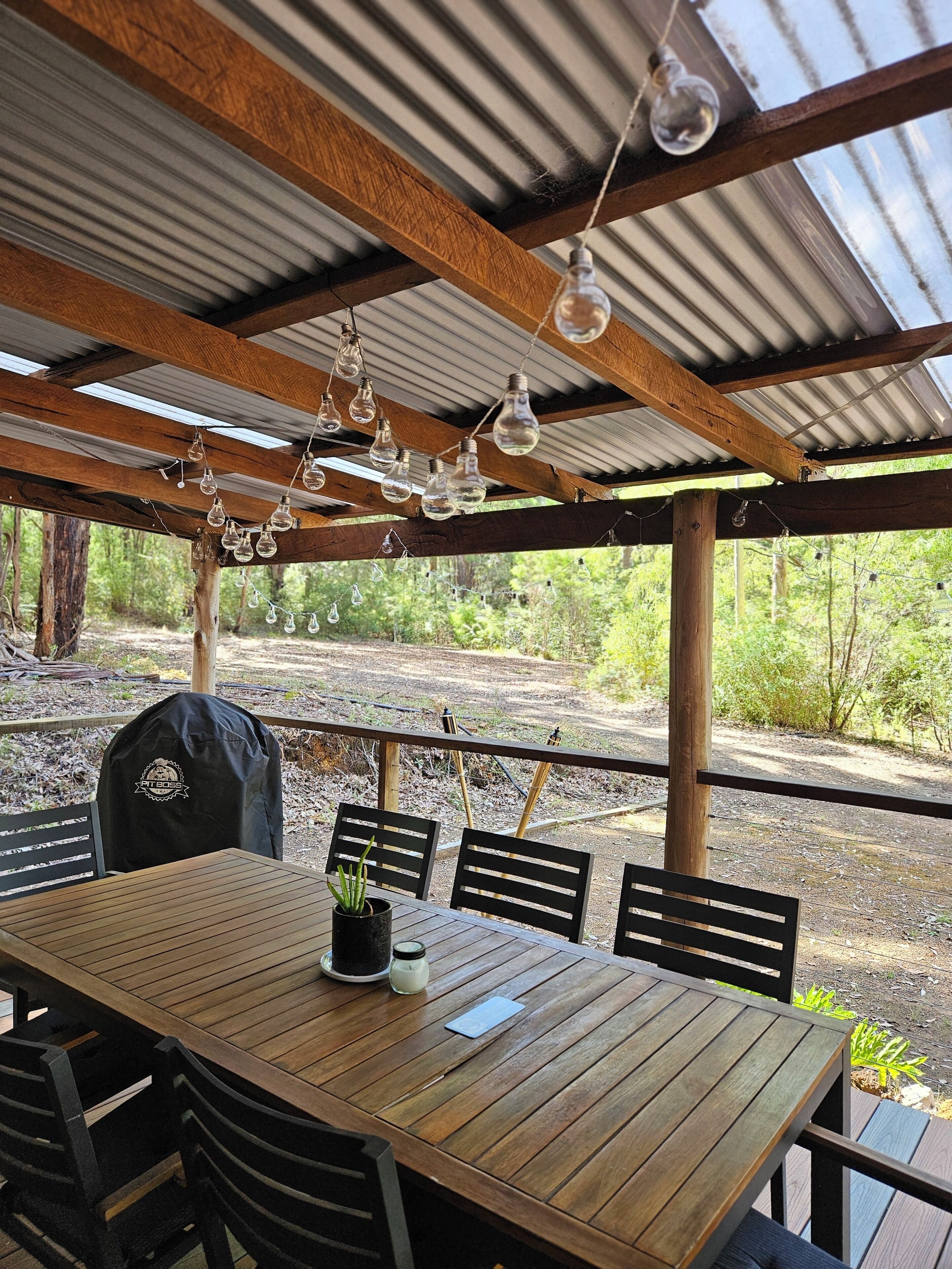 Een houten buitentafel met stoelen, een barbecue en hangende lampen onder een metalen overkapping.