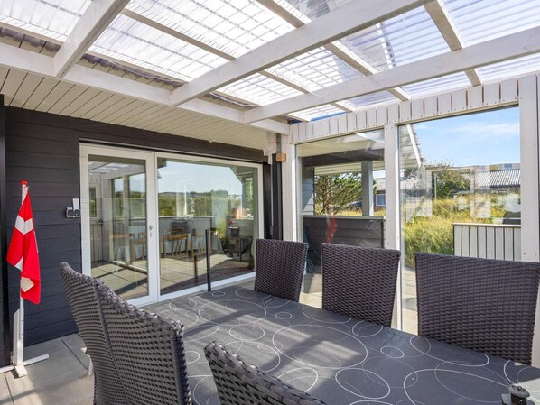 A covered outdoor patio with a table and chairs, glass doors leading to an interior space, and a flag on the left. - 4 Star Holiday Home in Norre Nebel (Norre Nebel)