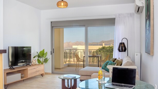 A modern living room with a flat-screen TV, a laptop on a glass table, a sofa with cushions, a potted plant, and a sliding glass door leading to a balcony with a view of mountains.