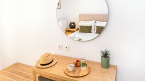 A bedroom with a round mirror, a wooden bedside table with a straw hat, a glass of water, a red flower, and a small green plant.