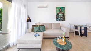 A modern living room with a beige sofa, a glass coffee table, and a dining table with chairs. There is a painting on the wall and an air conditioning unit above the sofa.