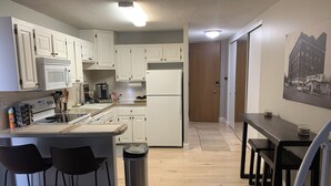 Eine moderne Küche mit weißen Schränken, einem Edelstahlkühlschrank, einer Mikrowelle und einer Kaffeemaschine. Zwei Barhocker und ein Mülleimer sind ebenfalls vorhanden.