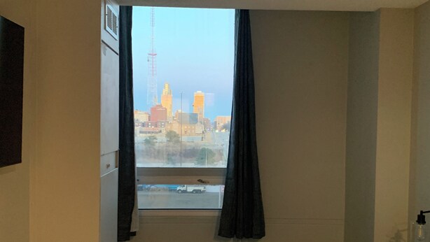 Ein Hotelzimmer mit einem großen Bett, einem Fenster mit Vorhängen, Blick auf eine Stadtlandschaft und einem an der Wand montierten Fernseher.