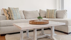 A modern living room with a light-colored sectional sofa, patterned throw pillows, a round woven coffee table, and a small potted plant.