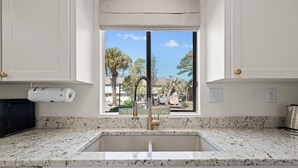 A kitchen with a granite countertop, a stainless steel sink, and a window with a view of palm trees and buildings.