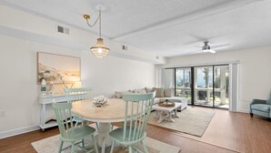 A spacious dining area with a round table, light green chairs, and a chandelier. There is a sofa and a coffee table in the background, and a sliding glass door leading to a balcony.