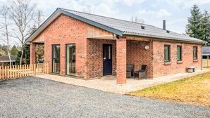 Une maison de plain-pied en briques, avec un toit en pente, un porche à l’avant doté d’un banc et une cheminée visible.