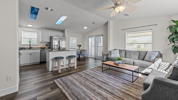 A spacious living area with a grey sofa, a coffee table, and a kitchen area with white cabinets and a central island with stools.