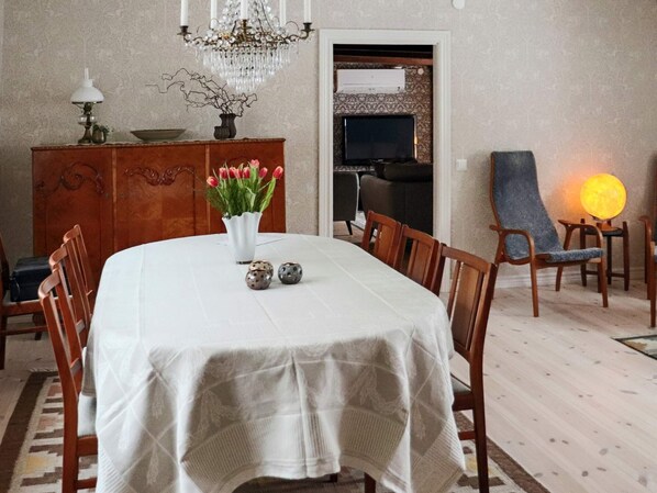 A dining area with a round table covered by a white tablecloth, surrounded by wooden chairs. A vase with red flowers and two decorative eggs are on the table. A chandelier hangs above, and a television is mounted on the wall. - 4 Star Holiday Home in Ullared (Ullared)