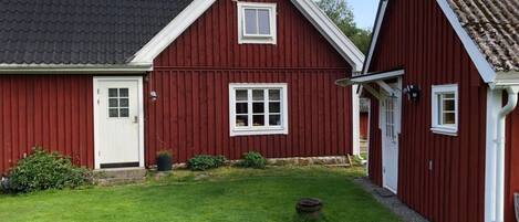Ein rotes Holzhaus mit weißer Tür und Fenstern, einem kleinen Garten und einem Schornstein.