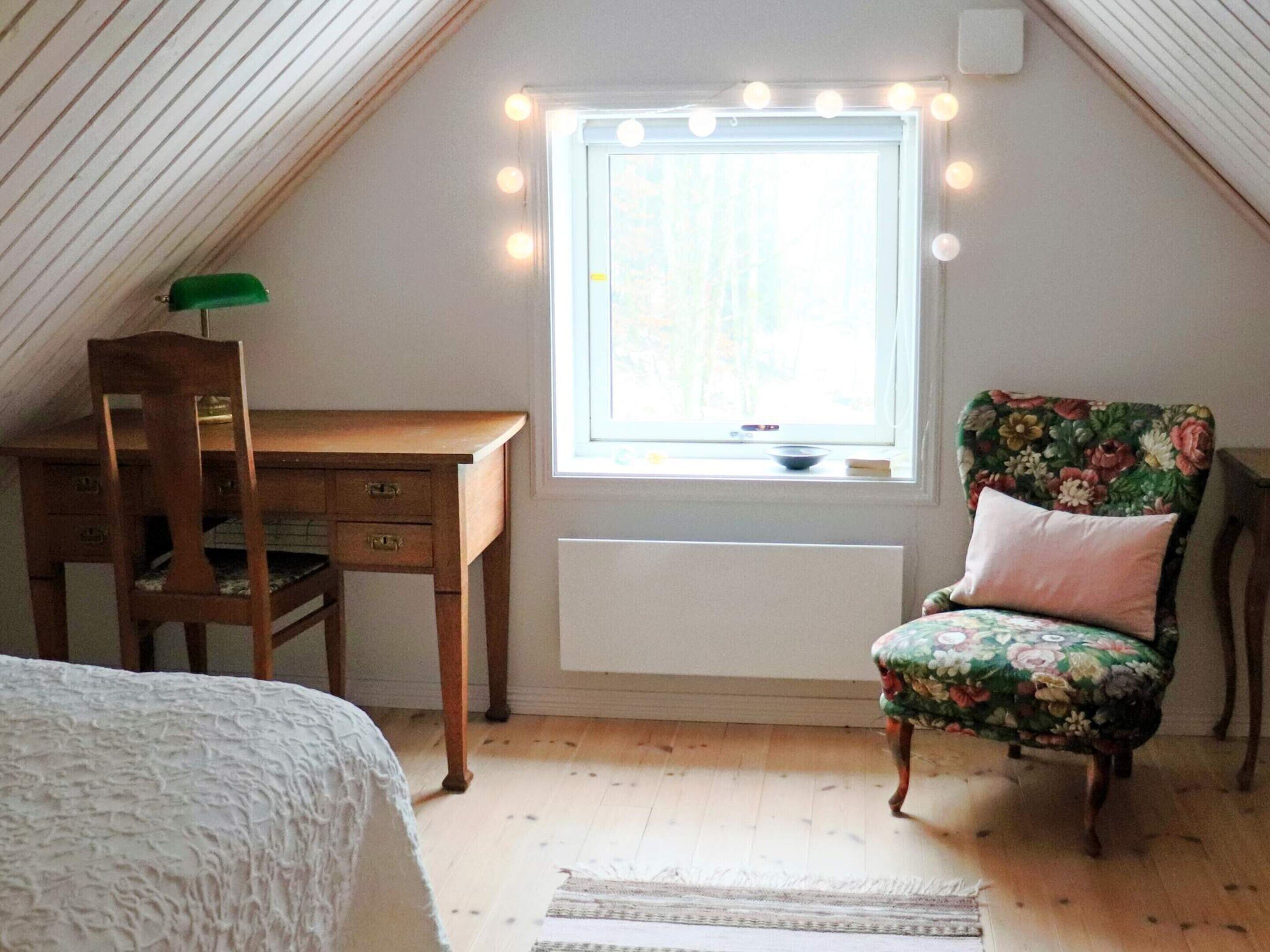 Ein gemütliches Schlafzimmer im Dachgeschoss mit schrägen Wänden, einem Holztisch, einem blumigen Sessel und einem großen Fenster mit Beleuchtung.