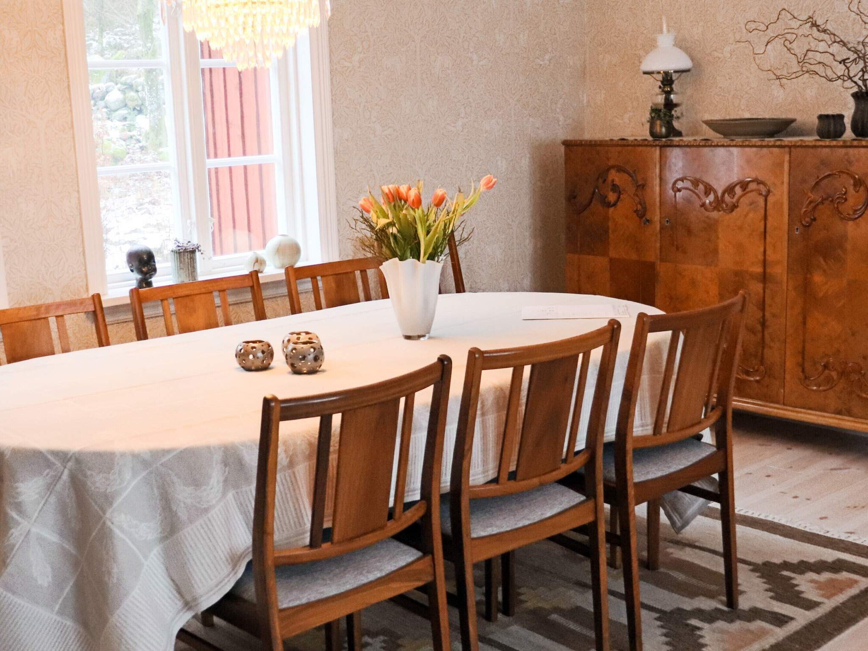 Een eetkamer met een ronde tafel, houten stoelen, een witte tafelkleed, een vaas met bloemen en een houten kast met een lamp.