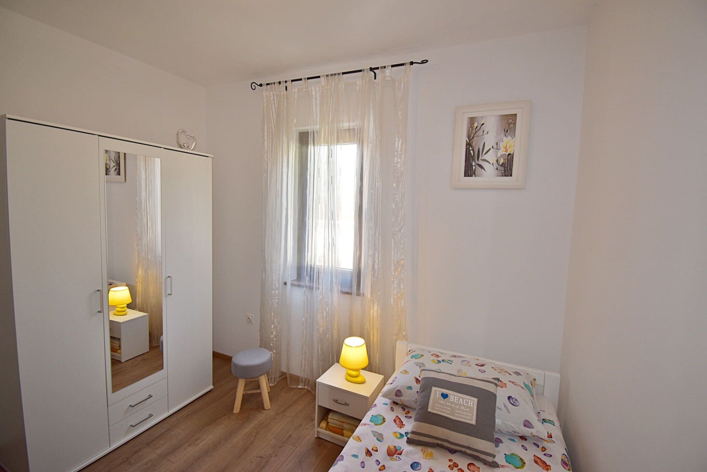 A bedroom with a bed, a nightstand, a chair, a wardrobe, and a window with curtains.