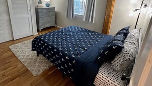 A bedroom with a wooden ceiling, a bed with navy blue bedding, a dresser, a window with curtains, and a floor lamp. - 3-bedroom beachy apartment one block from Hampton Beach (Hampton)