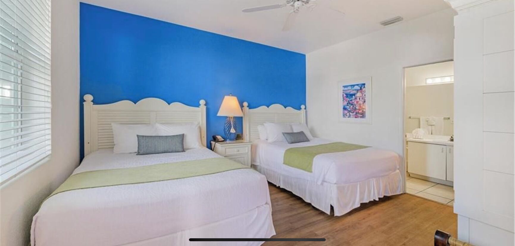 A hotel room with two beds, a blue accent wall, a ceiling fan, and a bathroom visible through an open door.