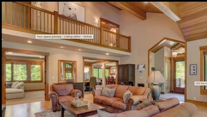 A spacious living room with a high ceiling, wooden beams, and large windows. There is a dining area and a kitchen visible in the background.