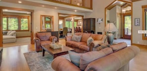 A spacious living room with a high ceiling, wooden beams, and large windows. There is a dining area and a kitchen visible in the background.