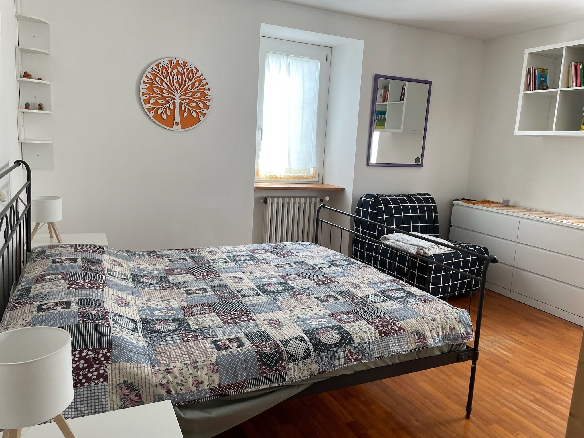Ein Schlafzimmer mit Bett, Nachttisch, Lampe, Heizkörper, Fenster mit Vorhängen, Wanddeko, Spiegel und Bücherregal.