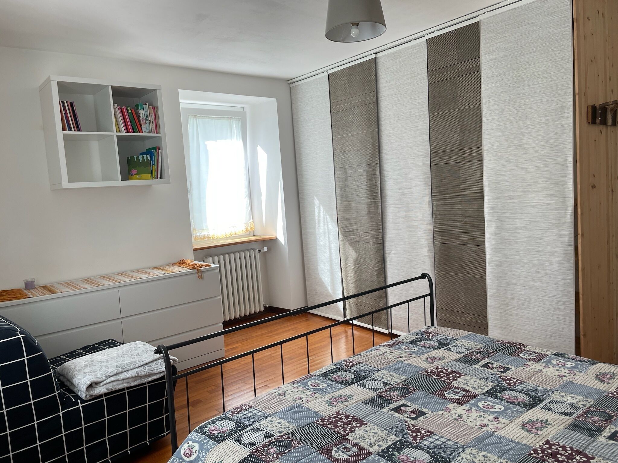 Ein Schlafzimmer mit Bett, Fenster, Heizkörper, Bücherregal und Kleiderschrank.