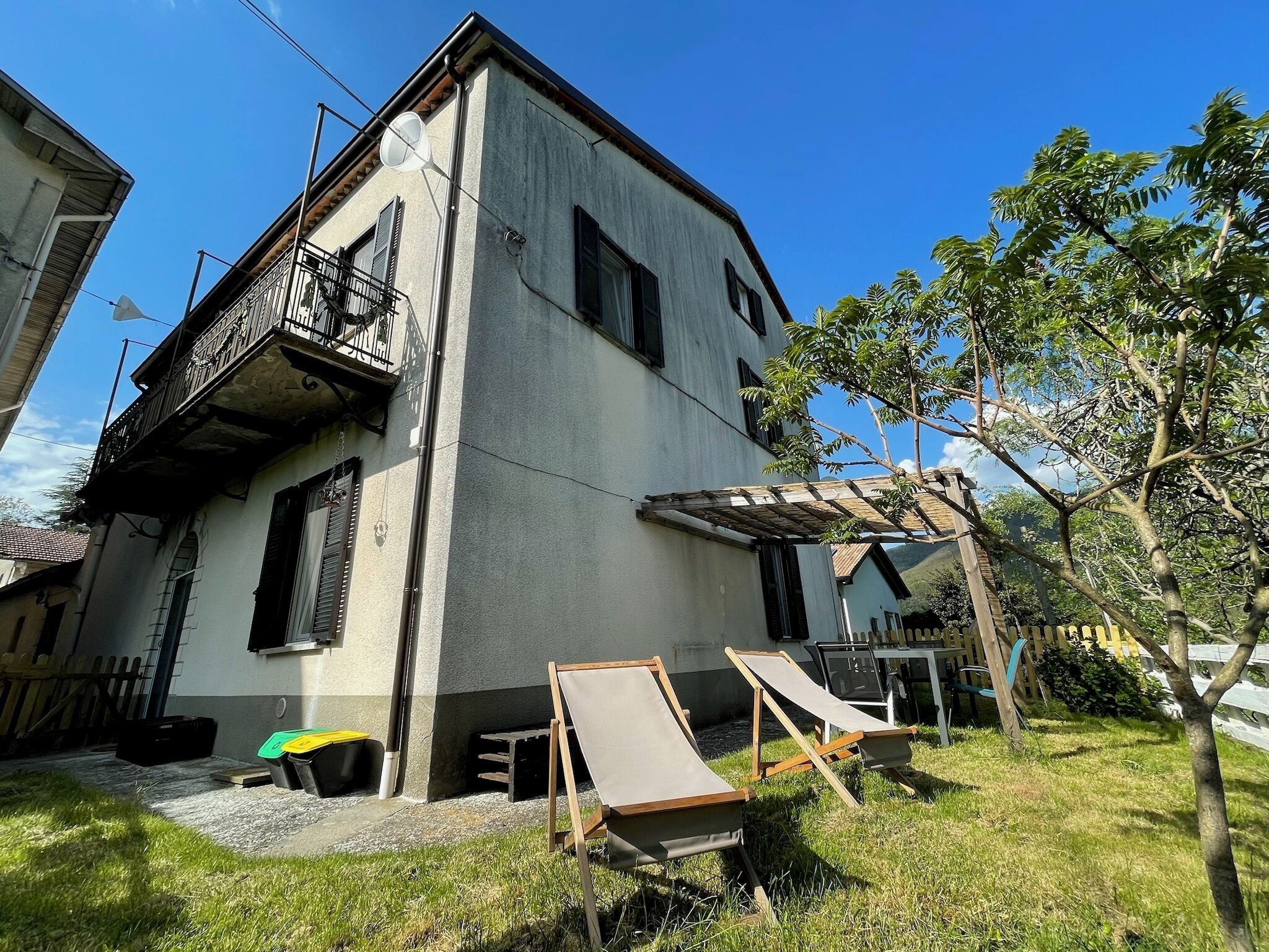 Uma casa de dois andares com uma varanda, um pequeno jardim com cadeiras de grama e um cercado de madeira.