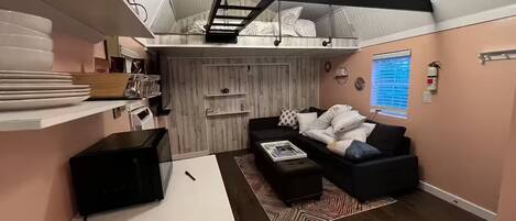Ett kompakt bostadsutrymme med en sovplats på ett loft, en soffa, ett soffbord och ett köksområde med mikrovågsugn.
