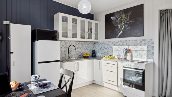 Cocina compacta con armarios blancos, microondas, estufa y fregadero. Hay una mesa de comedor con una computadora portátil y un plato de muffins.