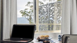 Una computadora portátil sobre una mesa con un plato de muffins y una taza, cerca de una ventana con vistas a árboles y montañas.