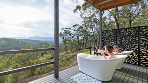 Una pareja relajándose en una bañera blanca sobre una terraza de madera, con una vista panorámica de un bosque y montañas.