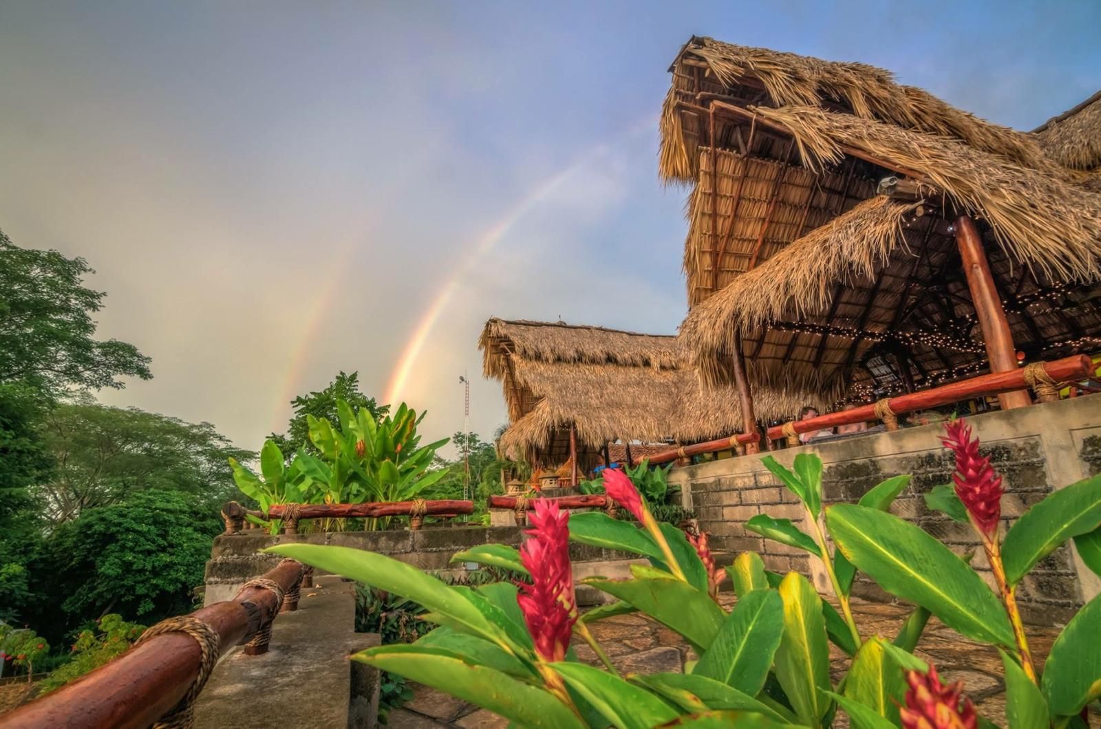 A thatched-roof building with a staircase leading up to it, surrounded by greenery and a rainbow in the background.