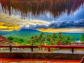 Una vista panoramica da un balcone con ringhiera, su un paesaggio rigoglioso con alberi e una catena montuosa in lontananza.