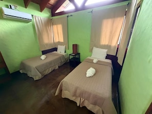 A room with two single beds, each with a pillow and a towel, a small table, and an air conditioning unit on the wall.