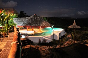 Vista nocturna de un resort con piscina, una estructura de techo de paja y un sendero de piedra.