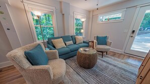 A modern living room with a sofa, two armchairs, a coffee table, and a large window with a view of greenery. - Charming Coastal Oasis with Heated Pool in Kitty Hawk, NC (Kitty Hawk)