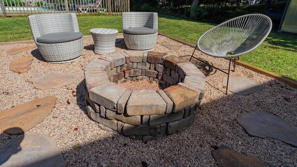 A circular stone fire pit surrounded by outdoor furniture, including two wicker chairs and a metal chair, set on a gravel surface with a wooden deck in the background. - Charming Coastal Oasis with Heated Pool in Kitty Hawk, NC (Kitty Hawk)