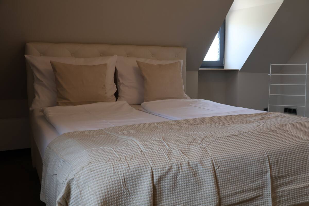 Een netjes opgemaakt bed met een hoofdbord, een raam met uitzicht en een witte muur.
