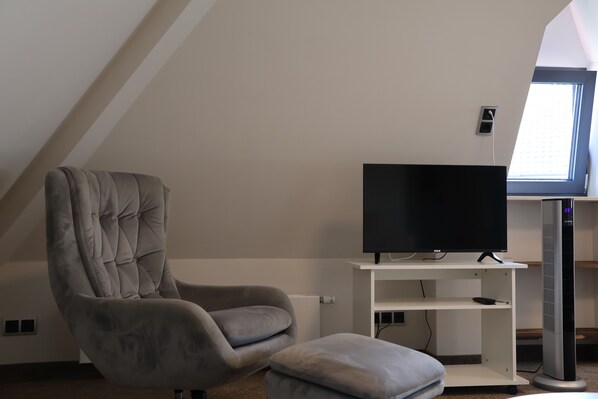 A modern living room with a high ceiling, a comfortable grey armchair, a flat-screen TV on a white stand, and a large window providing natural light. - Hotel an der Therme (Bad Durkheim)